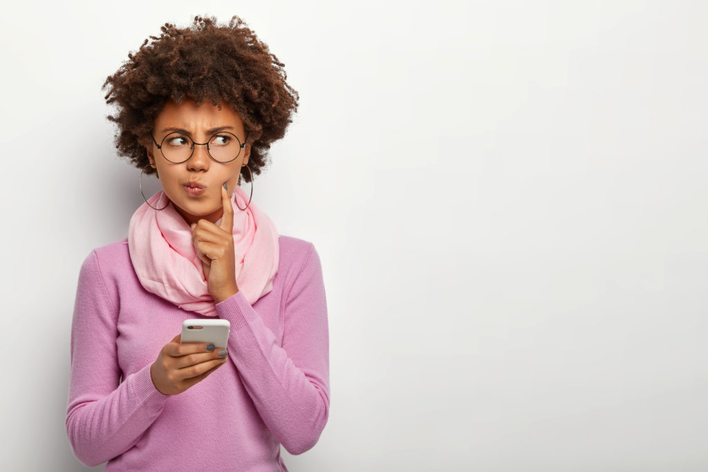 Pensive Dark Skinned Woman Looks Thoughtfully Aside, Holds Mobile Phone, Waits For Call, Purses Lips, Has Afro Haircut, Dressed In Violet Jumper With Scarf, Keeps Index Finger On Cheek, Poses Indoor - OFFICE CONT - Contabilidade Digital para você e sua empresa