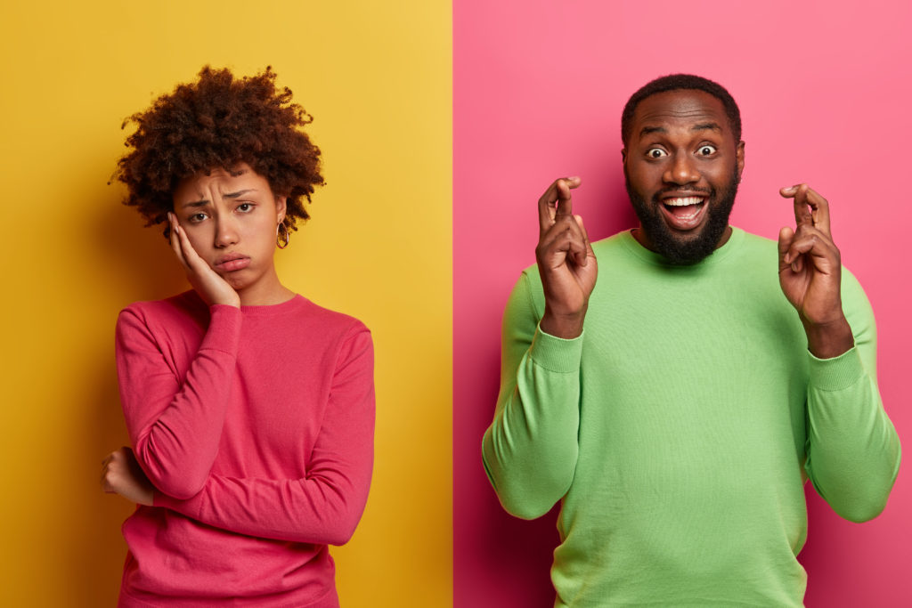 Upset Fatigue Curly Woman Looks Sadly At Camera, Her Boyfriend Stands Happy Near, Keeps Fingers Crossed, Believes In Good Fortune, Wears Green Jumper, Stand Against Yellow And Pink Background - OFFICE CONT - Contabilidade Digital para você e sua empresa